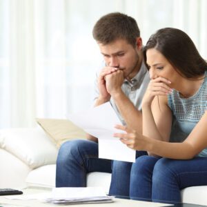 An image of a young couple sitting on a couch looking at a piece of paper with a worried expression on their faces