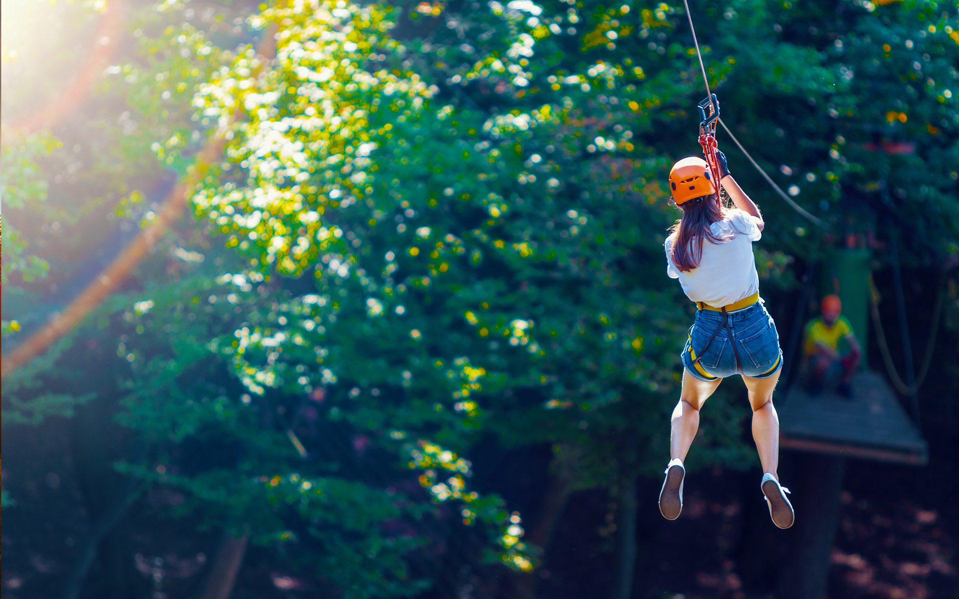 Zipline & Aerial Park Insurance Guide - Woman Ziplining Down to a Wooden Platform with People on a Sunny Day in a Forest